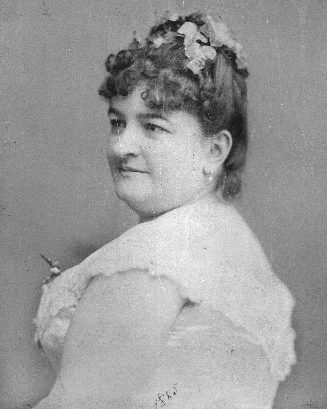 Fotografía antigua de Emilia Pardo Bazán, de medio perfil y mirando por encima del hombro, con vestido claro y flores en el pelo rizado